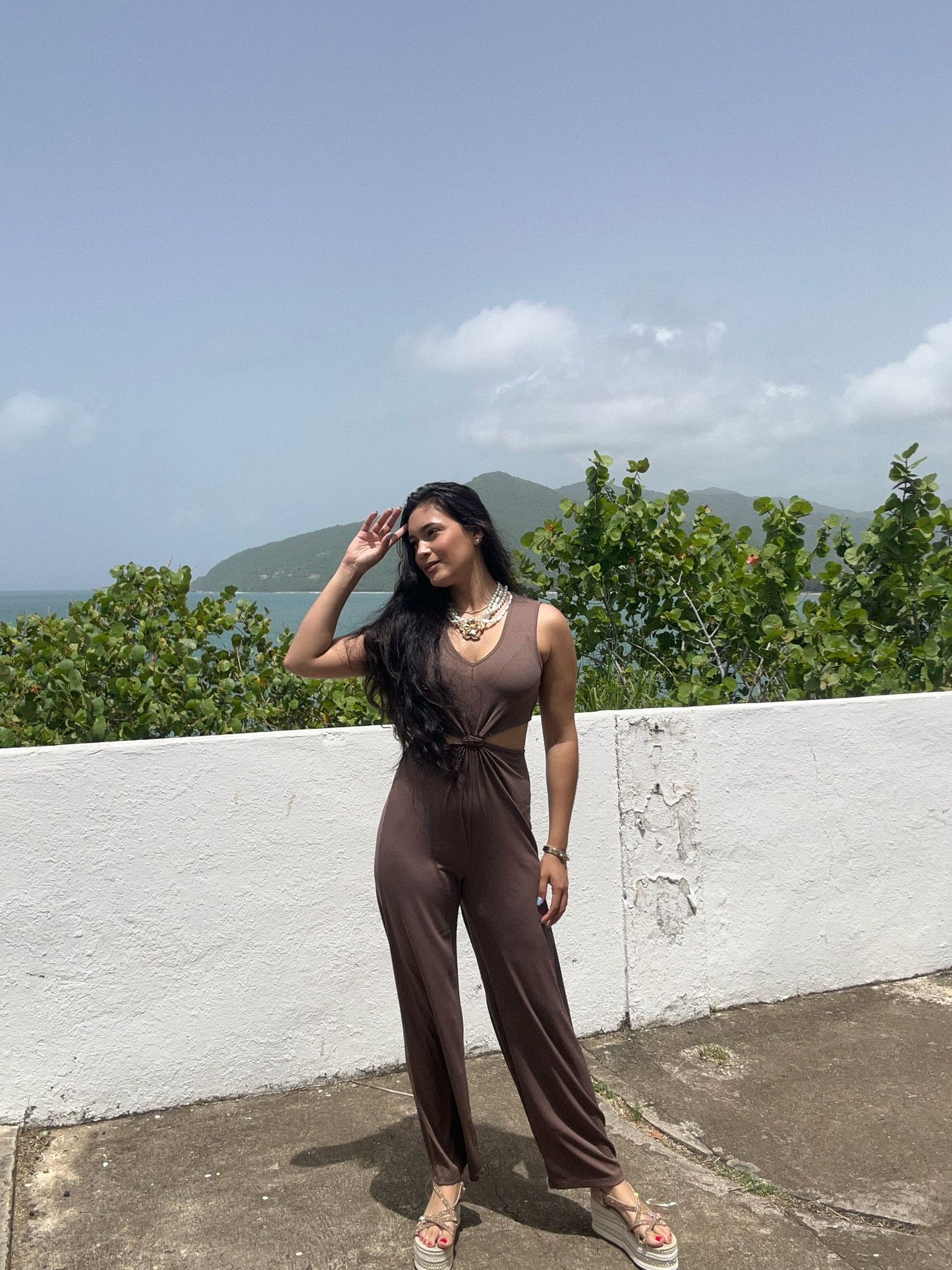 Woman in a brown jumpsuit standing outdoors with greenery and mountains in the background
