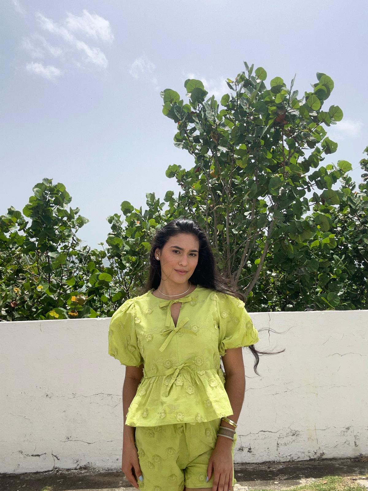 Woman wearing a bright green outfit standing outdoors with trees and a clear sky in the background