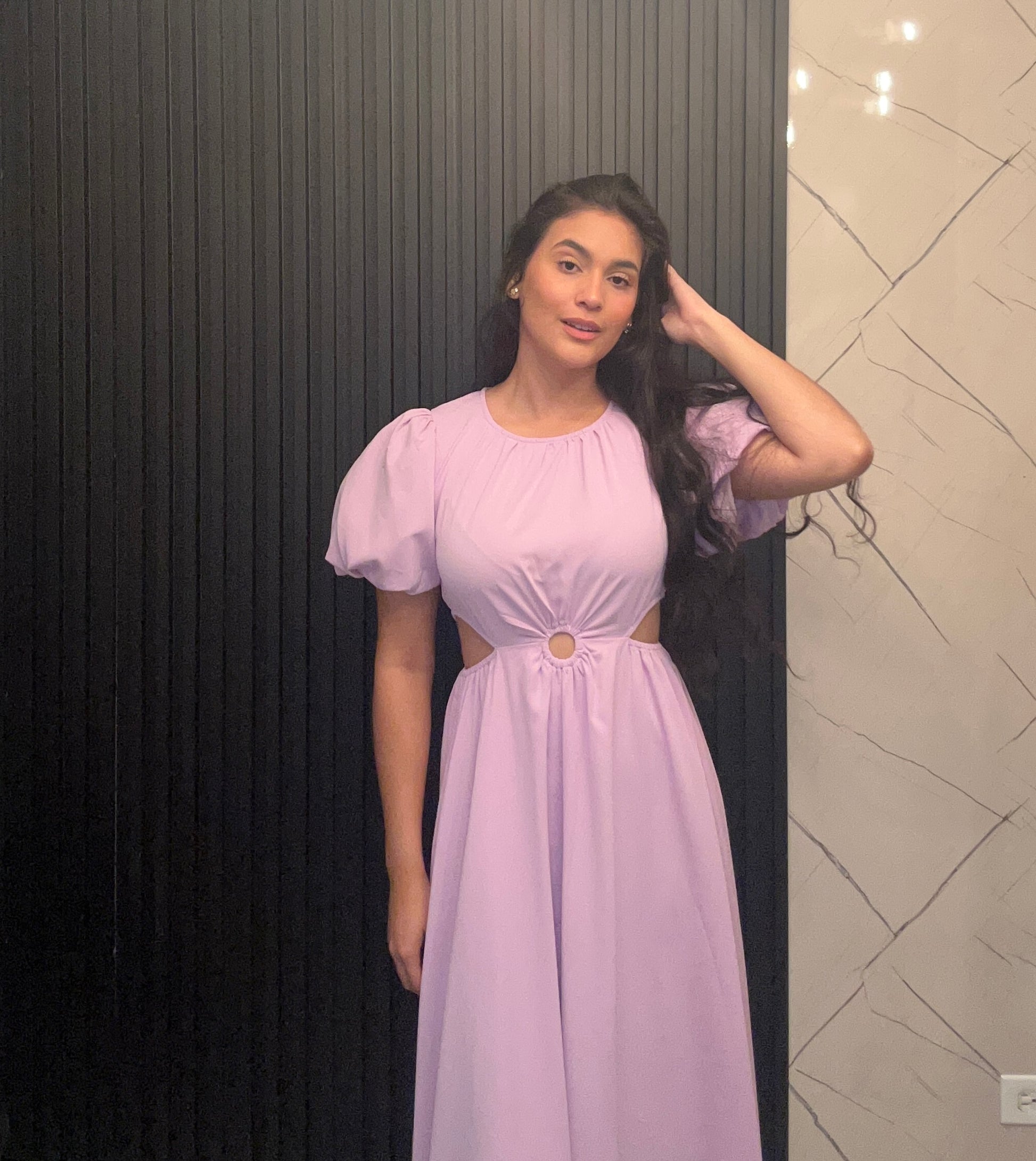 Woman wearing a light purple dress with puffed sleeves in front of a black and beige wall.