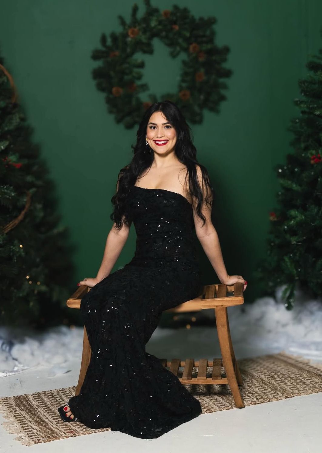 Woman in a black evening gown sitting on a wooden stool against a festive background with wreaths and trees.