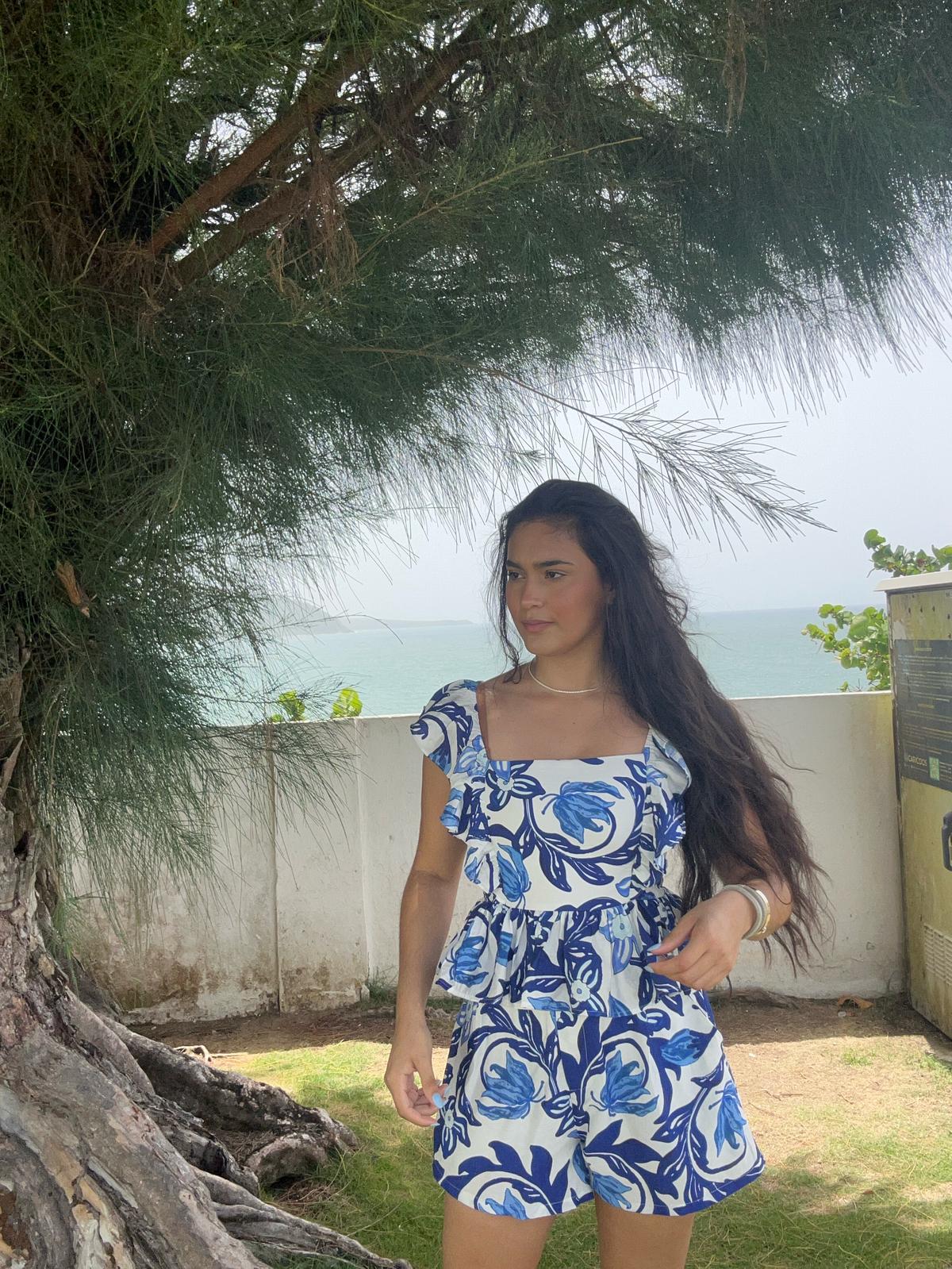Woman in a blue floral dress standing outdoors with trees and ocean in the background